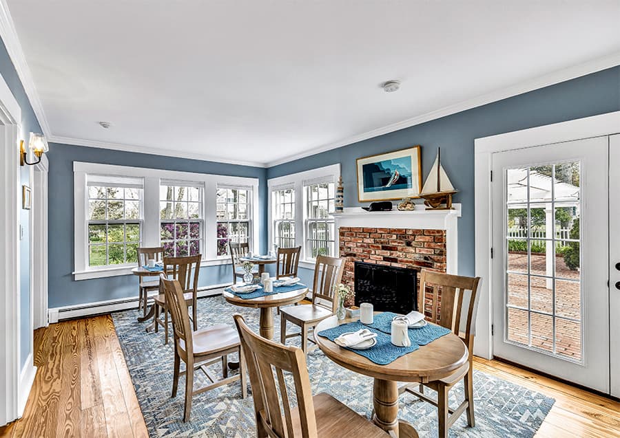 Bright dining area with wooden tables, chairs, a brick fireplace, and large windows.