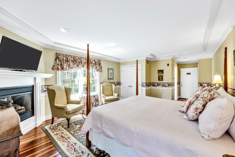A cozy bedroom featuring a king-size bed, fireplace, two armchairs, and large windows dressed in floral curtains.