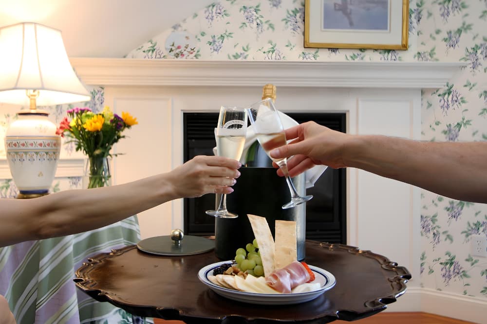Two hands clink champagne glasses over a platter of assorted cheeses, fruits, and snacks in a cozy, floral-decorated setting.